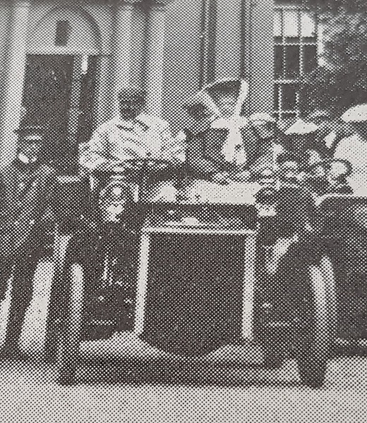 Historical black and white photograph of an early open motor car with two occupants